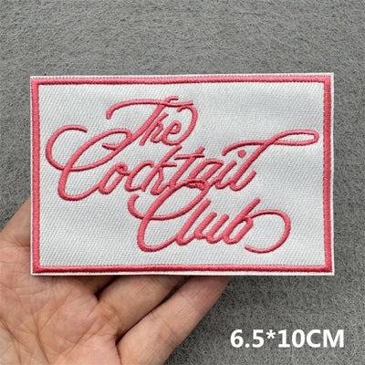 Embroidered patch with 'The Cocktail Club' text held by a hand on a gray background