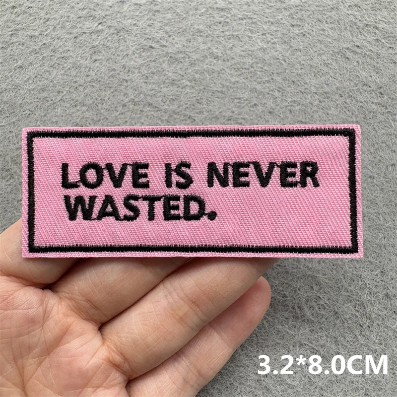 Pink patch with 'LOVE IS NEVER WASTED.' text held by a hand on a gray background
