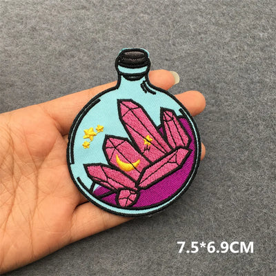 Embroidered patch of a bottle with pink crystals held by a hand on a gray background.