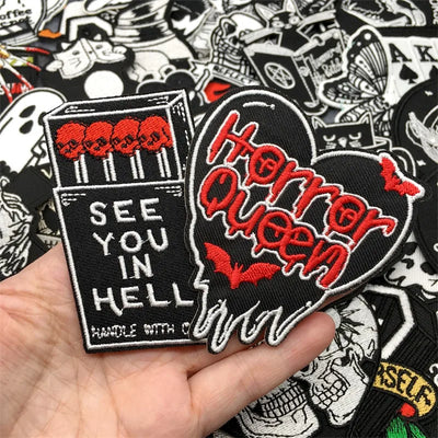 Hand holding a 'Horror Queen' patch with 'See You in Hell' text, surrounded by various other patches.