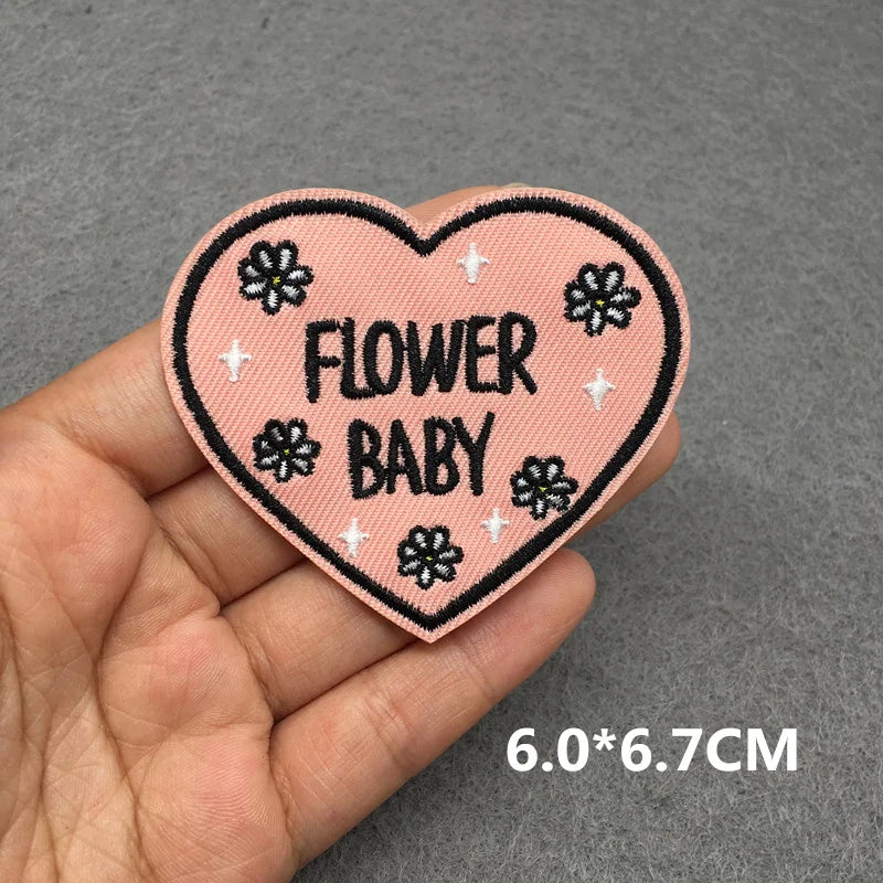 Heart-shaped patch with 'FLOWER BABY' text held in a hand on a gray background