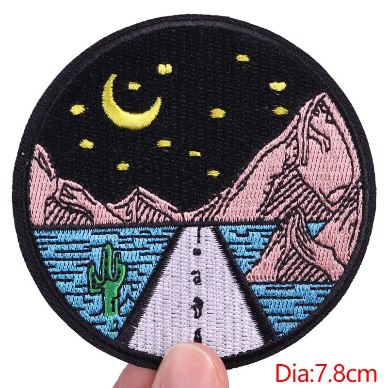 Embroidered patch with a night scene design held by a hand, showing a road, mountains, and moon.