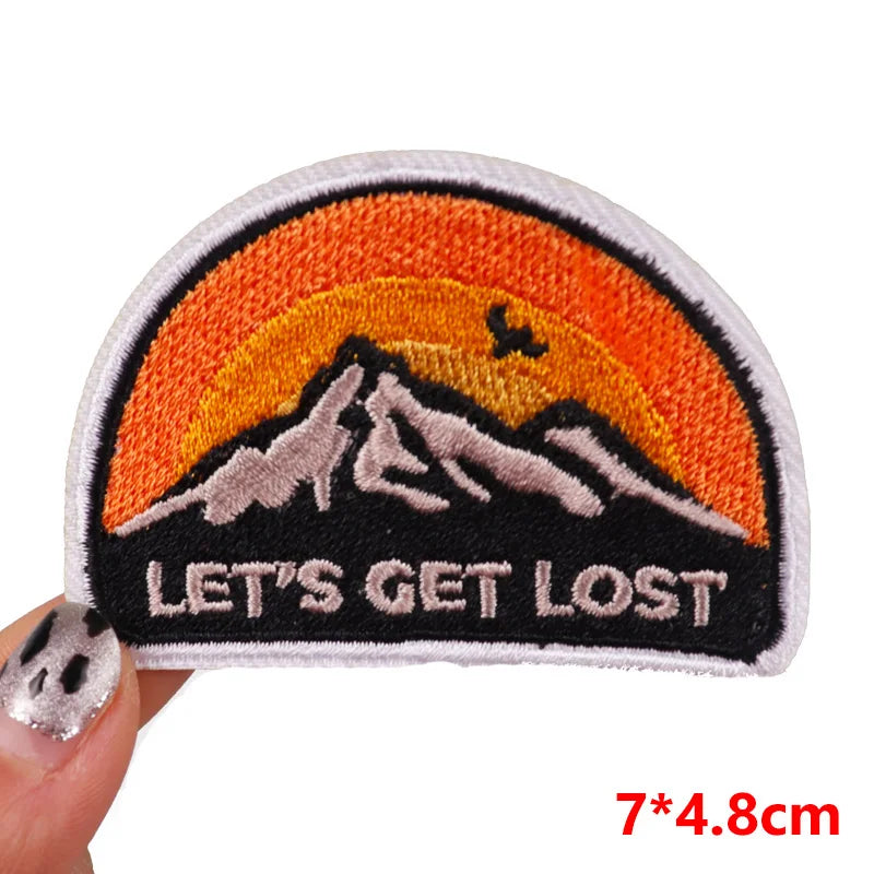 Embroidered patch with mountain design and 'Let's Get Lost' text held by a hand.