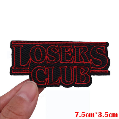 Embroidered patch with 'LOSERS CLUB' text held by a hand on a white background