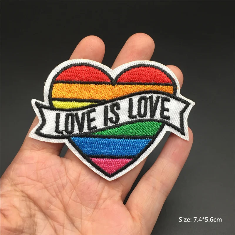 Heart-shaped patch with rainbow design and 'LOVE IS LOVE' text held in a hand against a dark background.