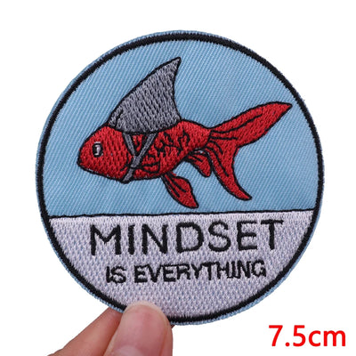 Circular patch with a red fish and 'Mindset is Everything' text, held by a hand against a white background.
