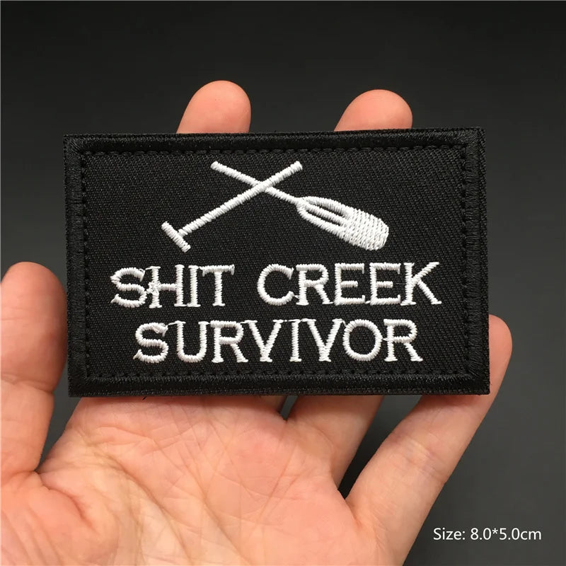 Black patch with 'SHIT CREEK SURVIVOR' text and oar design held in a hand against a dark background.