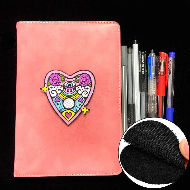 Pink notebook with a colorful heart-shaped patch and pens on a black background