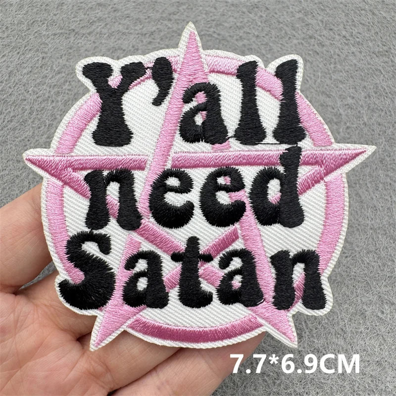 Embroidered patch with 'Y'all need Satan' text held by a hand on a gray background