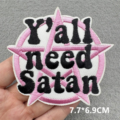 Embroidered patch with 'Y'all need Satan' text held by a hand on a gray background