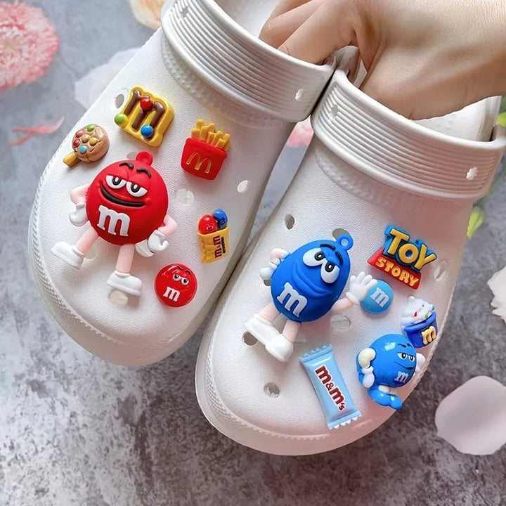 White clogs with colorful m&amp;m shoe charms a gray surface.