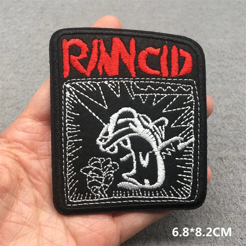 Hand holding a black red and white Rancid embroidered iron-on patch 6.8x8.2cm on a grey background