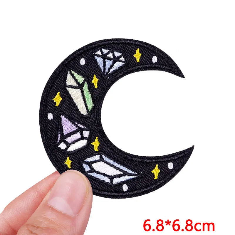 Black crescent moon patch with colorful crystals and stars held by a hand on a white background.