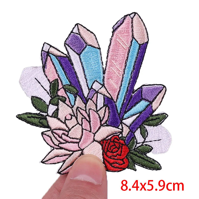 Embroidered patch with crystals, succulents, and a rose held by a finger on a white background.
