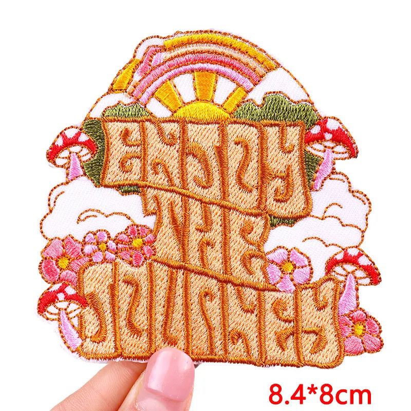 Embroidered patch with text, mushrooms, flowers, and a rainbow held by a hand on a white background.