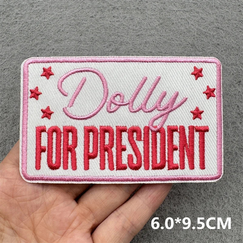 Hand holding a pink and white embroidered Dolly for President iron-on patch approximately 6x9.5cm on a gray background