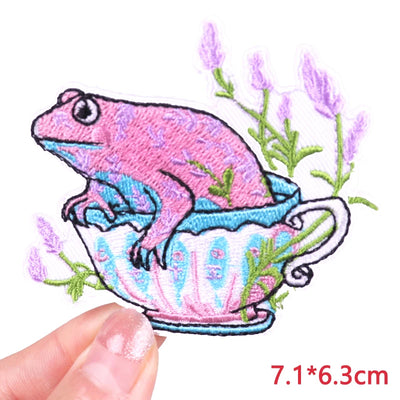 Embroidered patch of a pink frog in a teacup with lavender, held by a hand.