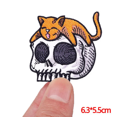 Embroidered patch of a cat on a skull design held by a finger, with dimensions displayed.