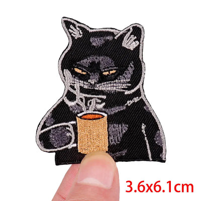Embroidered patch of a black cat holding a cup, held between a thumb and forefinger.