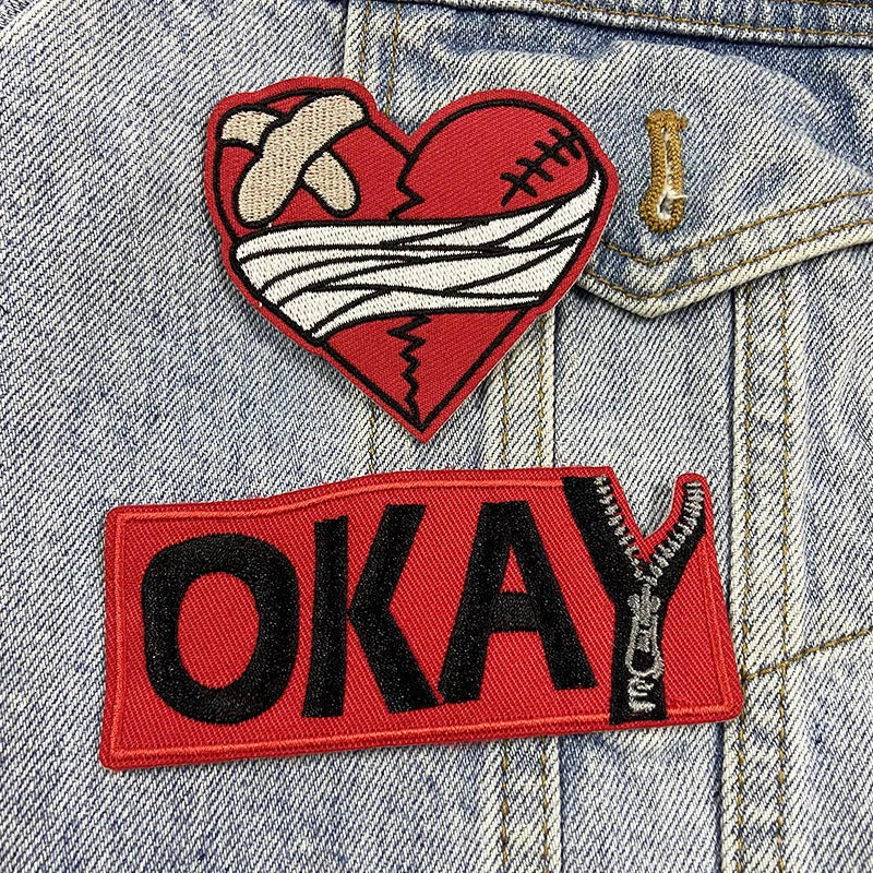 Two patches on a denim jacket: one heart-shaped with a bandage and the other 'OKAY' with a zipper.