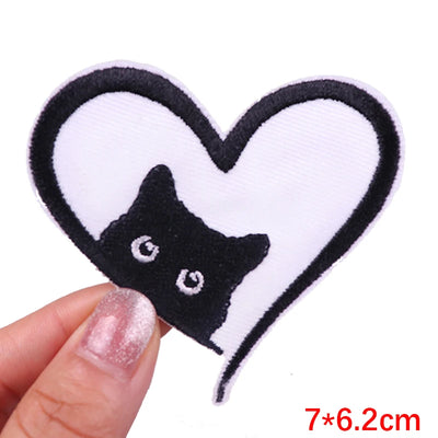 Heart-shaped patch with a black cat design held by a hand, on a white background.
