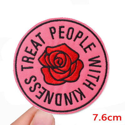 Pink patch with red rose and text 'Treat people with kindness' held by a hand on a white background.