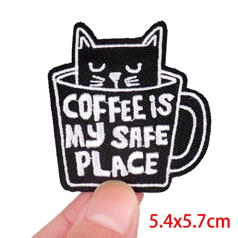 Embroidered patch with a cat in a coffee cup and 'Coffee is my safe place' text, held by a hand.