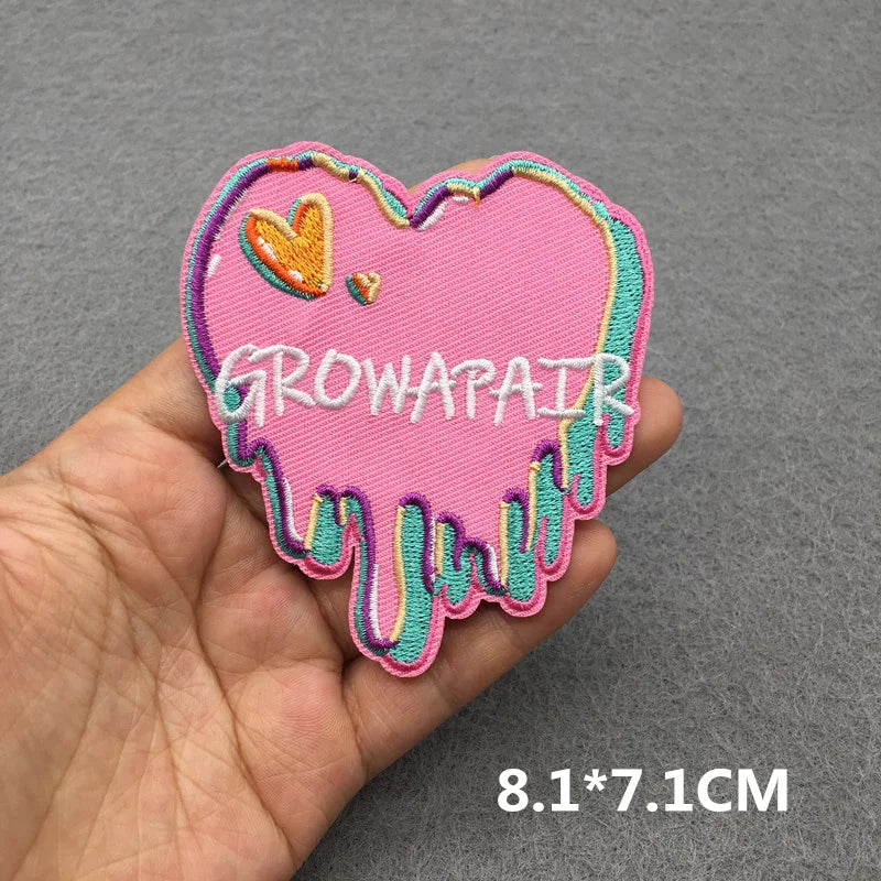 Pink heart-shaped patch with 'GROWAPAIR' text held in a hand against a gray background