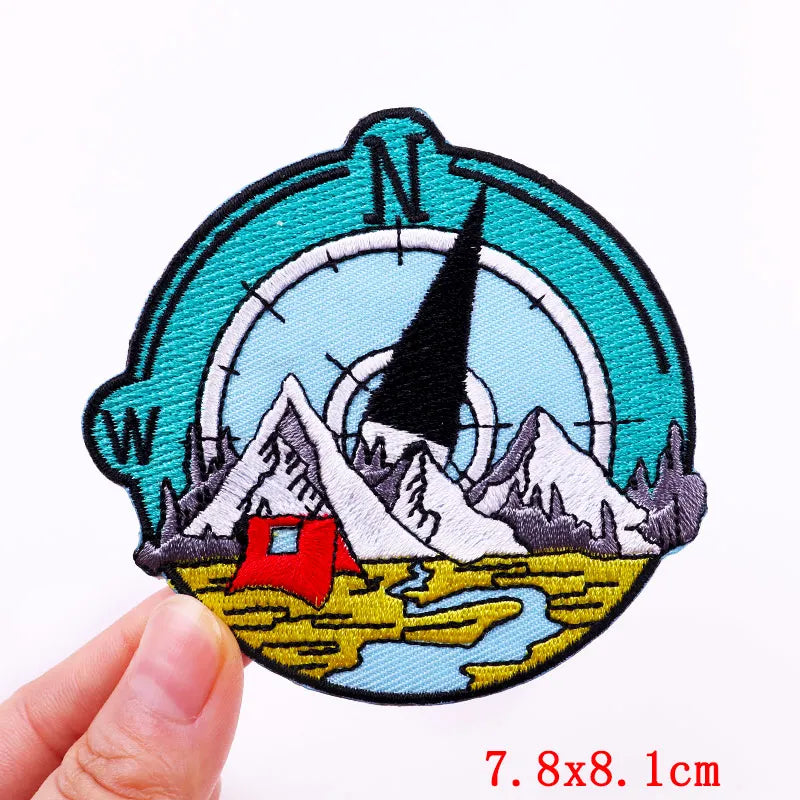 Embroidered patch with mountain and tent design held by a hand, on a white background.