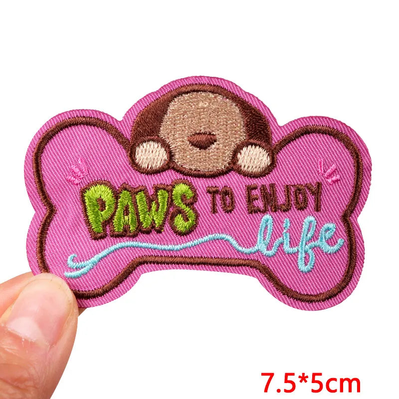 Pink bone-shaped patch with monkey design and text 'Paws to enjoy life' held by a hand.