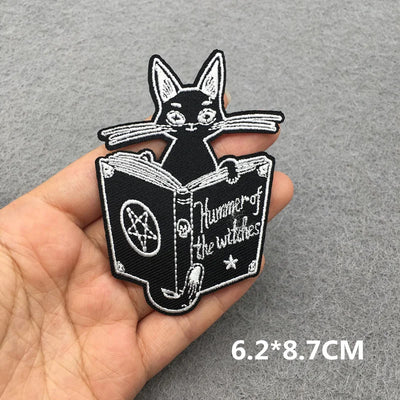 Embroidered patch of a black cat on an open book with text held in a hand against a gray background.