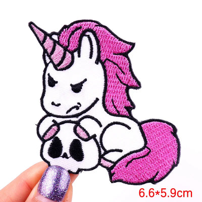 Embroidered patch of a unicorn with pink hair and a skull design, held by a hand with purple nail polish.