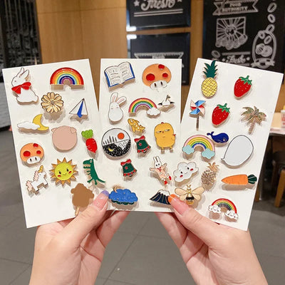 Three sheets of colorful enamel pins held by hands with a blurred indoor background.