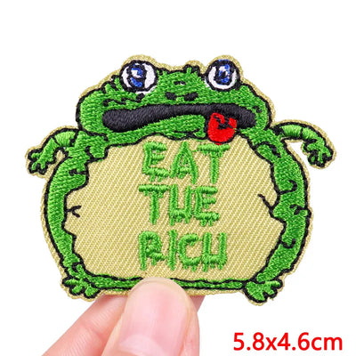 Embroidered patch of a frog with 'EAT THE FROG' text held by a hand on a white background.