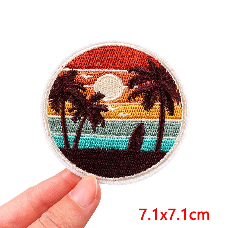 Circular embroidered patch of a sunset with palm trees held by a hand, on a white background.