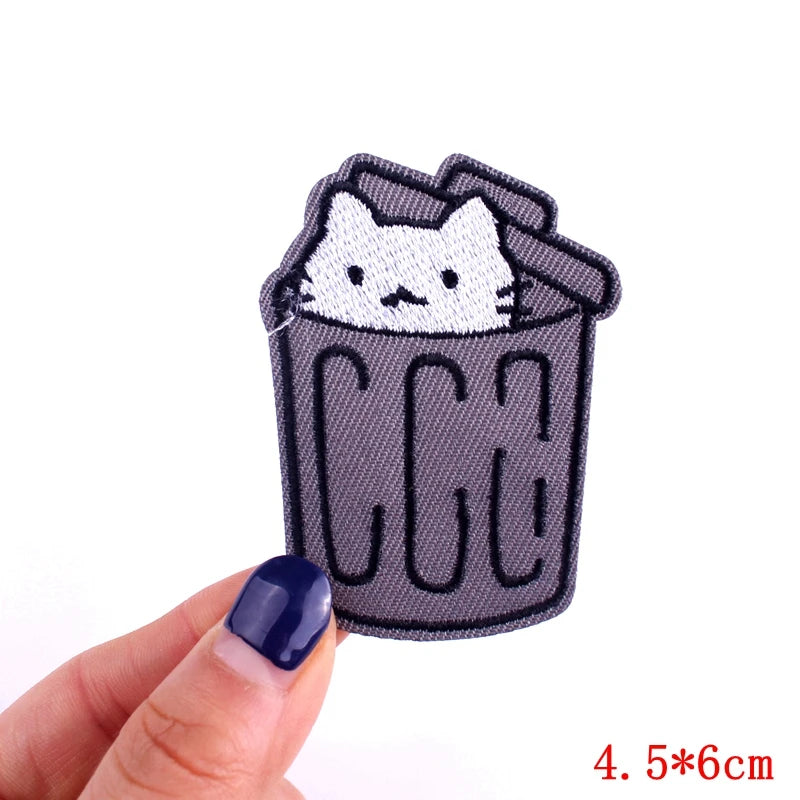 Patch of a cat in a trash can held by a hand with blue nail polish on a white background.