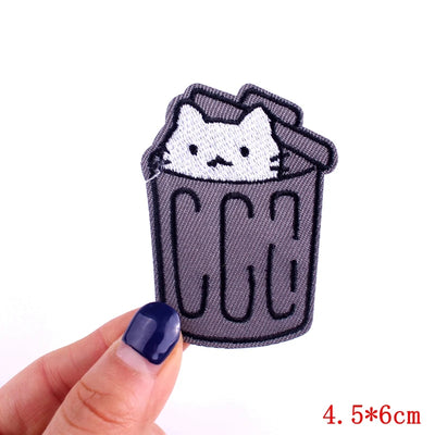 Patch of a cat in a trash can held by a hand with blue nail polish on a white background.