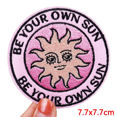 Circular patch with a sun illustration and text 'Be your own sun' on a pink background, held by a hand.