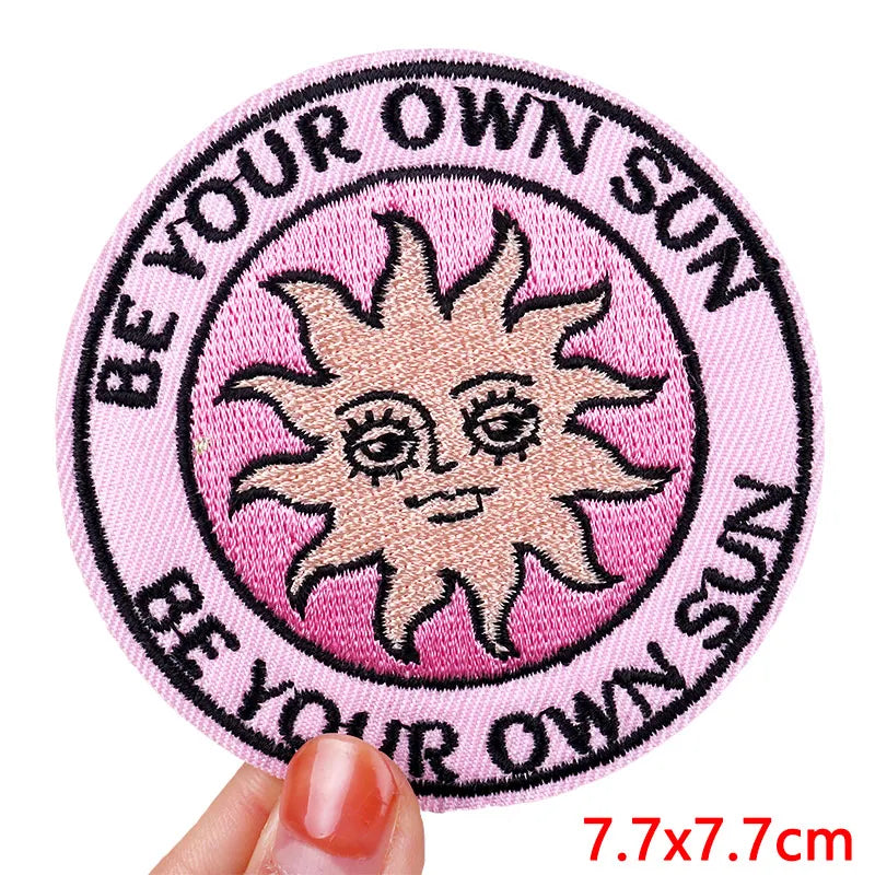 Circular patch with a sun design and text 'Be your own sun' on a pink background, held by a hand.