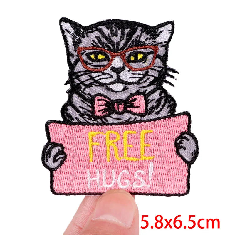 Embroidered patch of a cat holding a 'FREE HUGS!' sign, held between fingers with size indication.