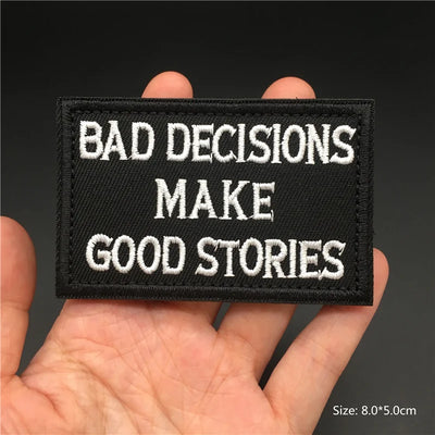 Black patch with white text 'BAD DECISIONS MAKE GOOD STORIES' held in a hand against a dark background.