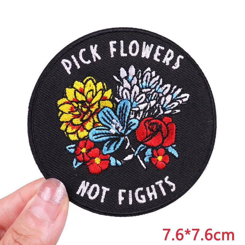 Round patch with floral design and text 'Pick Flowers Not Fights' held by a hand.