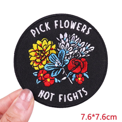 Round patch with floral design and text 'Pick Flowers Not Fights' held by a hand.