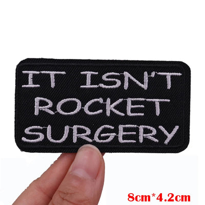 Embroidered patch with 'IT ISN'T ROCKET SURGERY' text held by a hand on a white background.