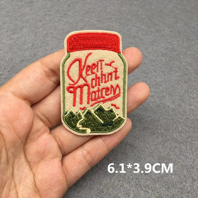 Embroidered patch of a can with 'Keen Munch Mates' text held in a hand on a gray background.