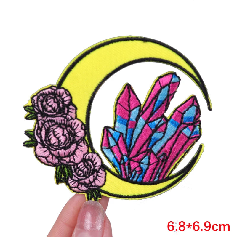 Embroidered patch with flowers and crystals on a yellow crescent moon, held by a hand.