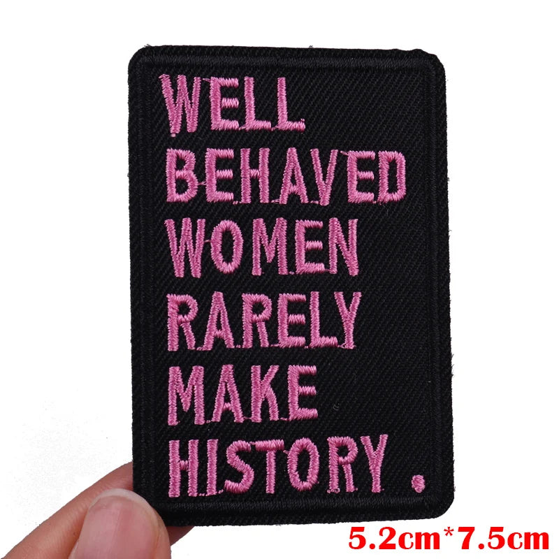 Black patch with pink text 'Well Behaved Women Rarely Make History' held by a hand on a white background.