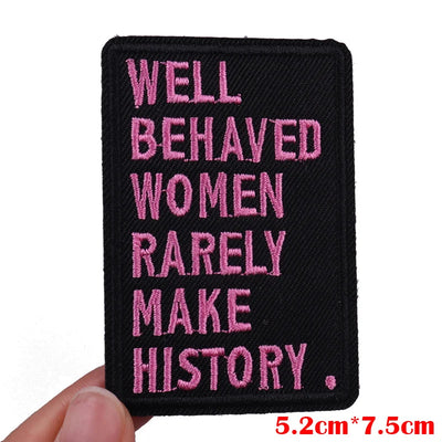 Black patch with pink text 'Well Behaved Women Rarely Make History' held by a hand on a white background.