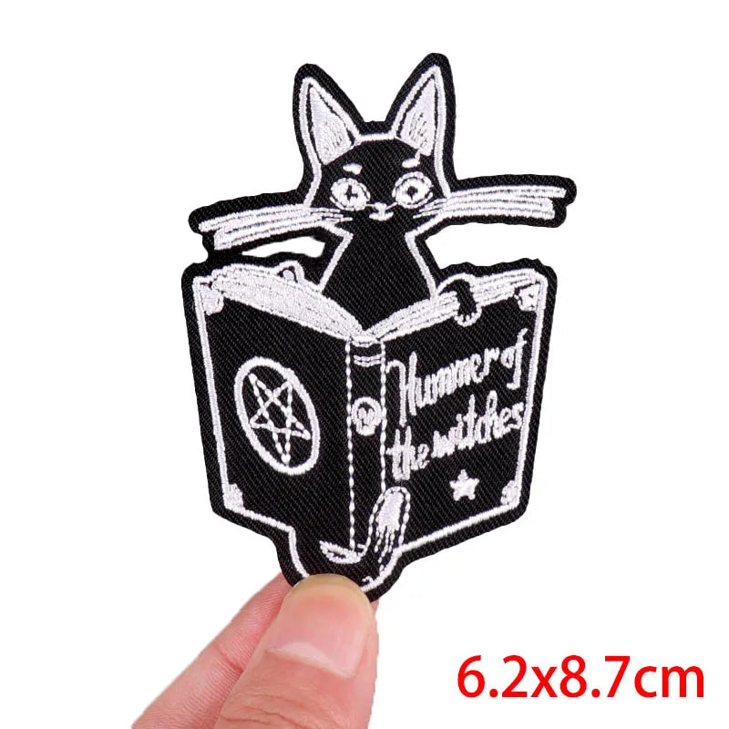 Embroidered patch of a cat reading a book titled 'Hunner of the Witches' held by a hand.