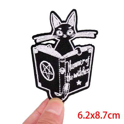 Embroidered patch of a cat reading a book titled 'Hunner of the Witches' held by a hand.